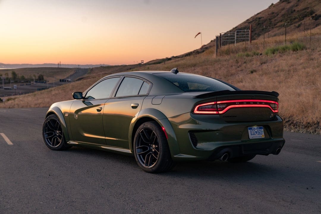 Rear view of a green Dodge Charger Scat Pack Widebody with glowing red tail lights on a road at sunset.