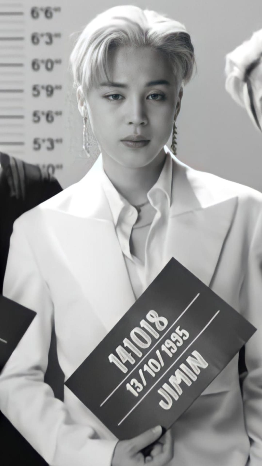 Jimin holds a police mugshot board in front of a height chart while wearing a white suit in monochrome.