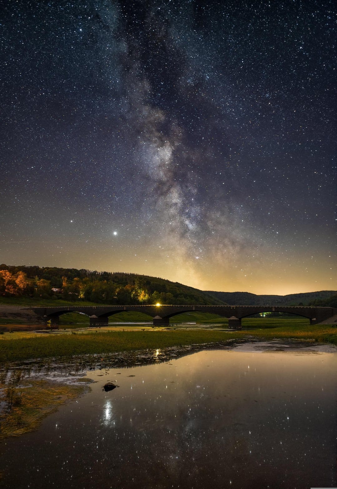 Milky Way galaxy arching over a stone bridge with a symmetrical reflection in a calm lake at night.