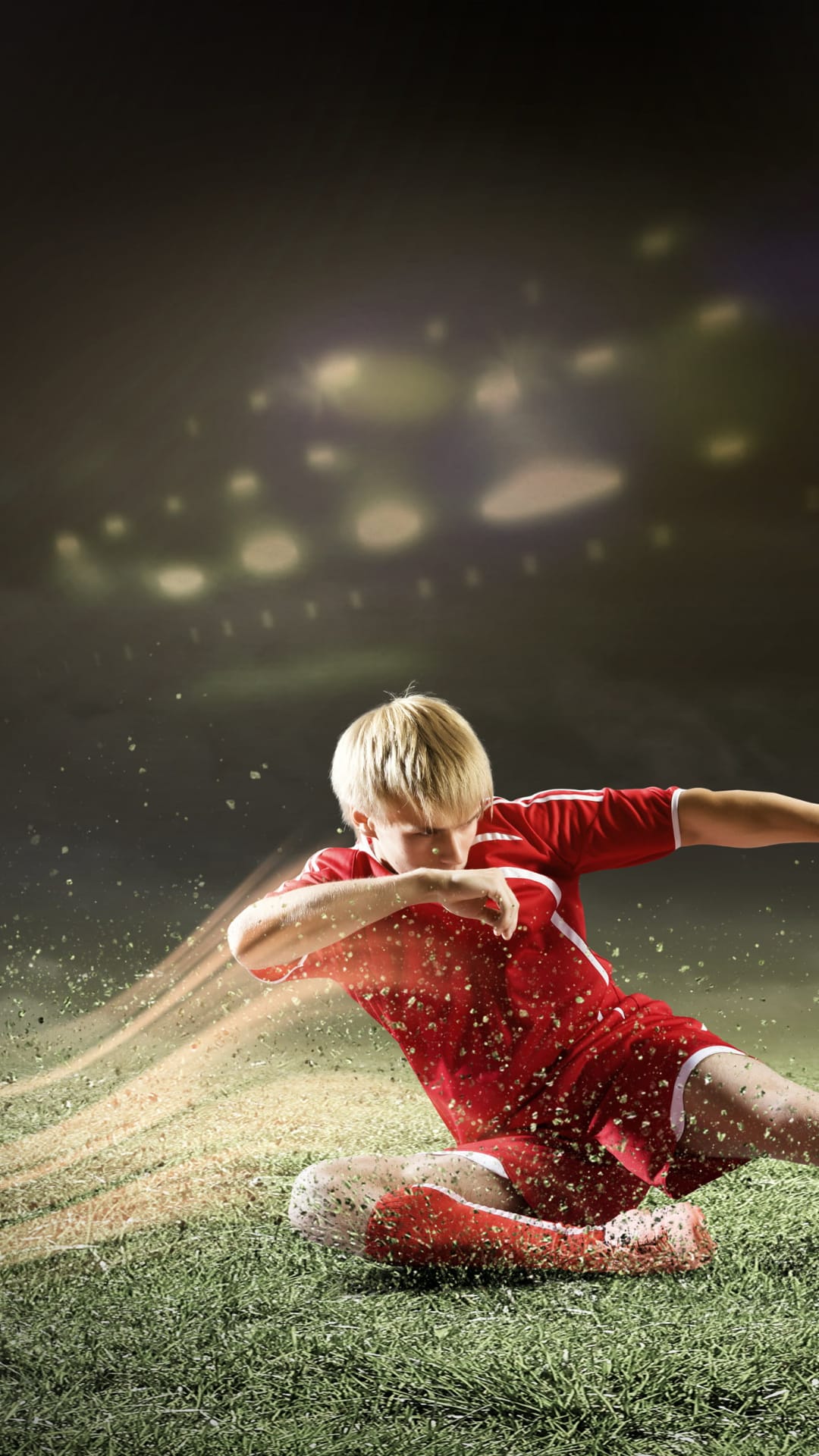 Young soccer player in a red jersey kicks a football with grass spraying under bright stadium floodlights.