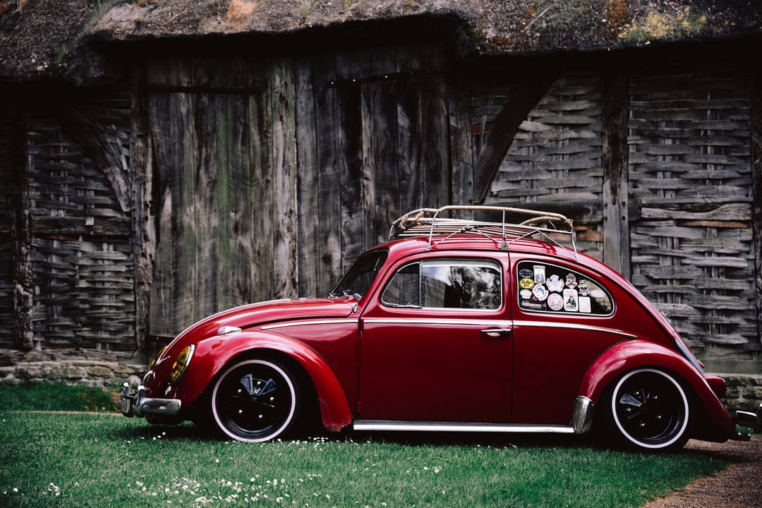 Vintage red Volkswagen Beetle with a roof rack parked on grass in front of a thatched-roof wooden building.