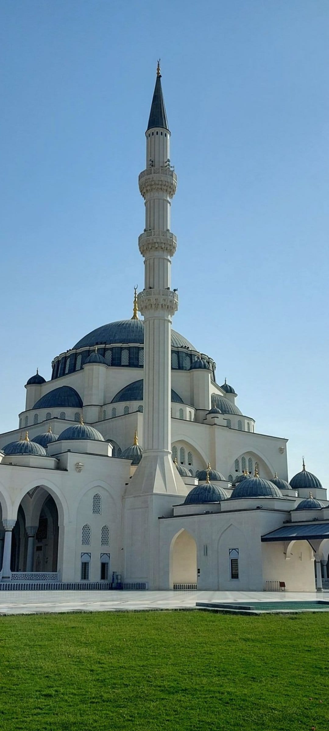White masjid featuring cerulean domes and a tall minaret overlooks a manicured green lawn under a bright blue sky.