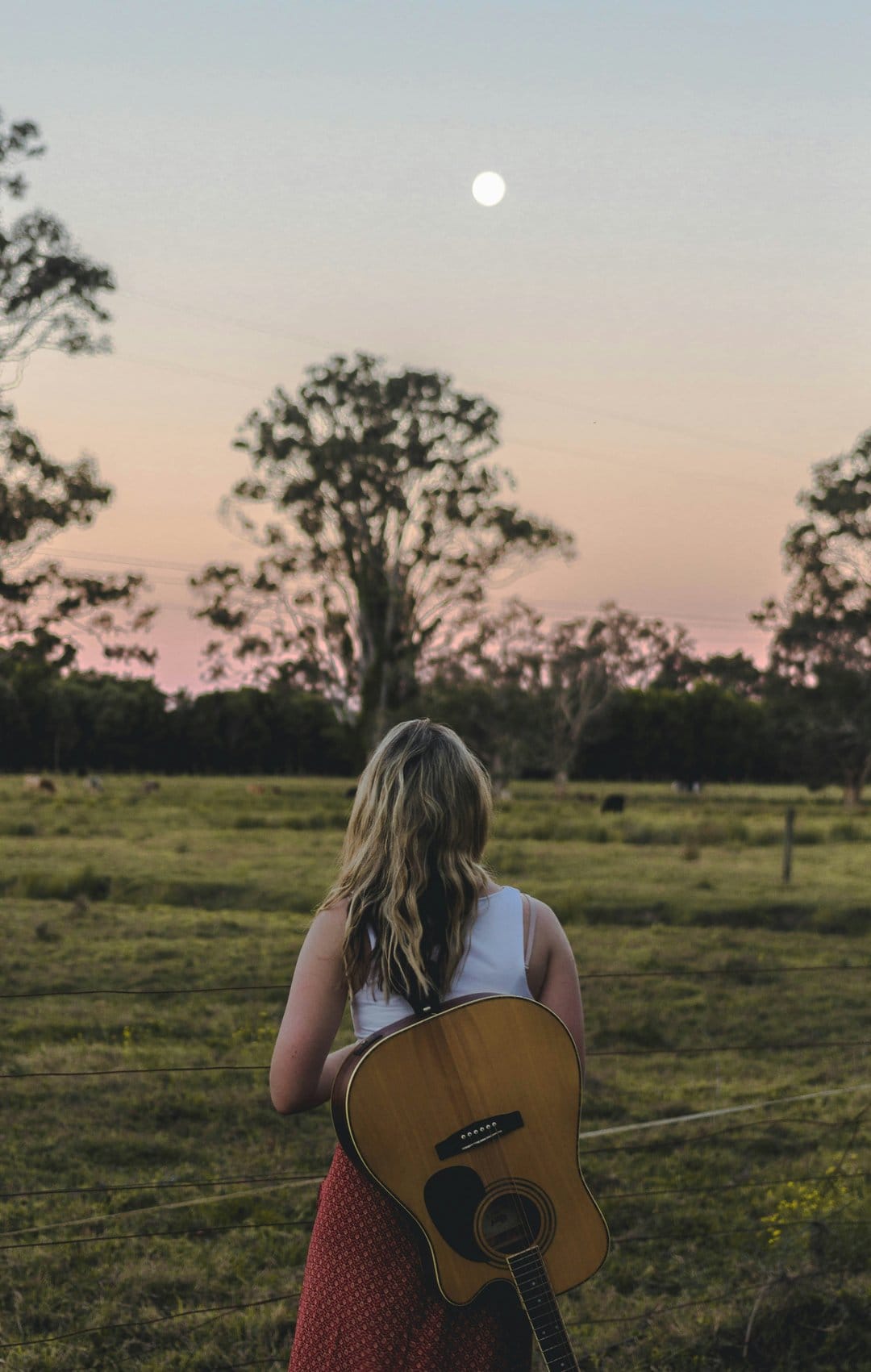 Woman holding a guitar faces a pastel sunset and full moon while standing in a grassy meadow during twilight.
