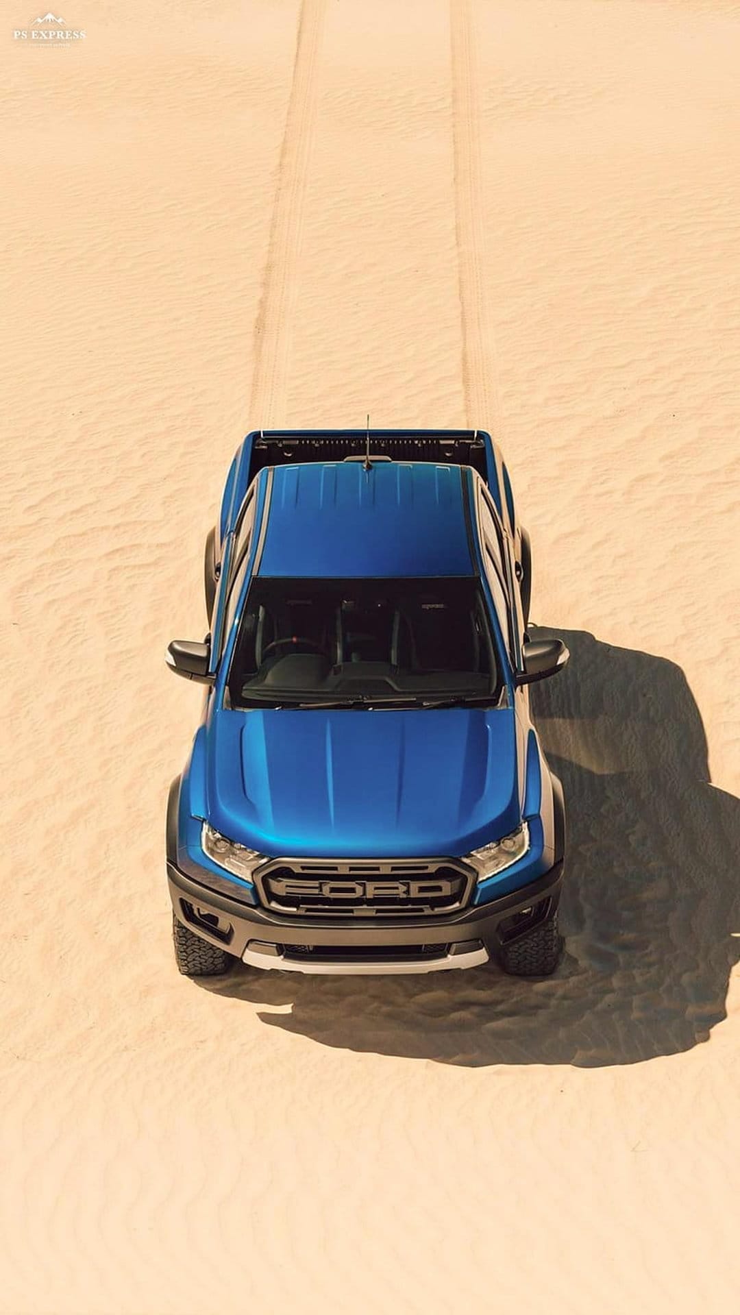 Blue Ford Ranger Raptor pickup truck drives across rippling desert sand dunes seen from a high aerial perspective.