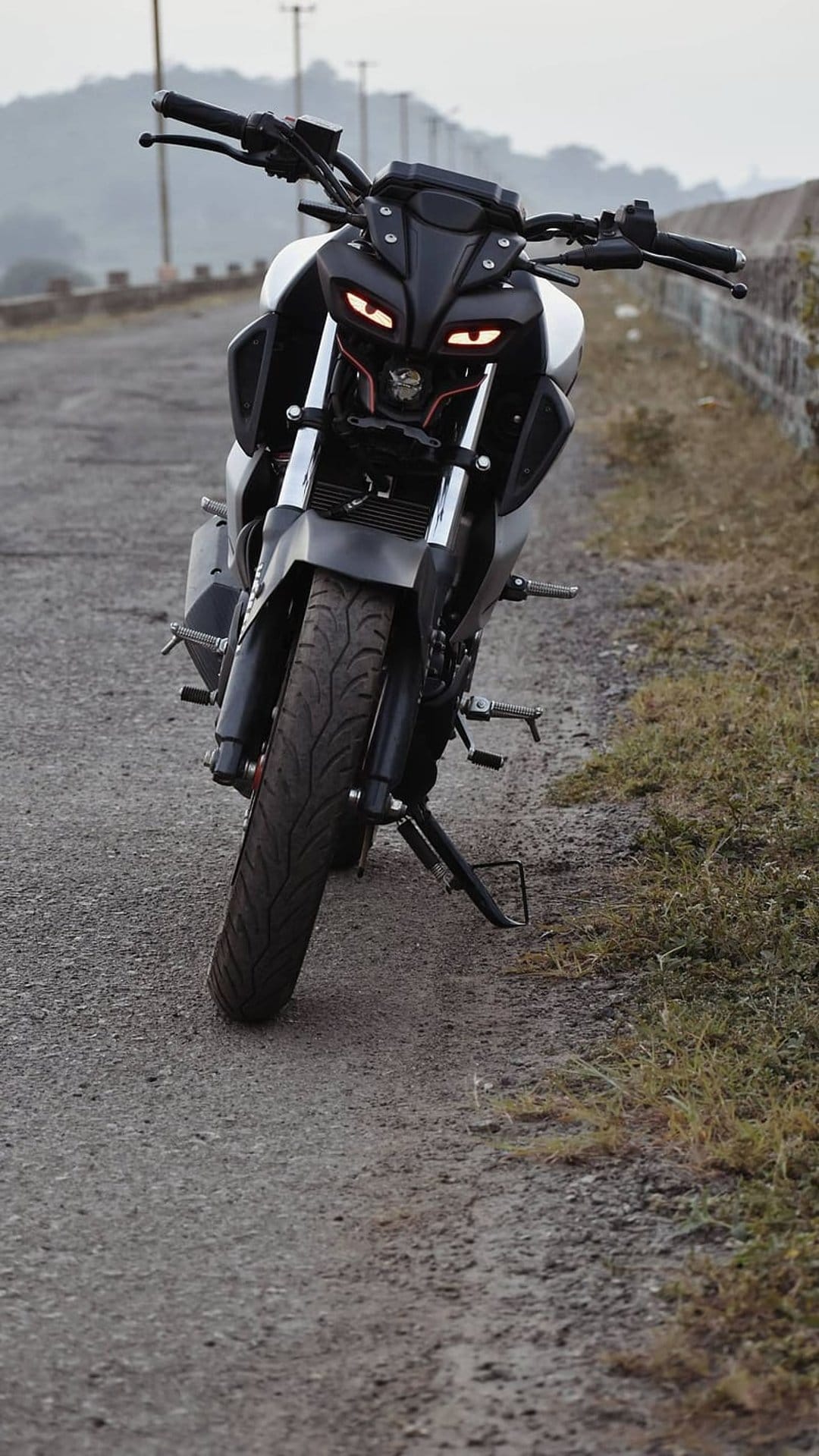 Yamaha MT-15 motorcycle with glowing orange LED lights faces forward on a mountain road under a hazy sky.