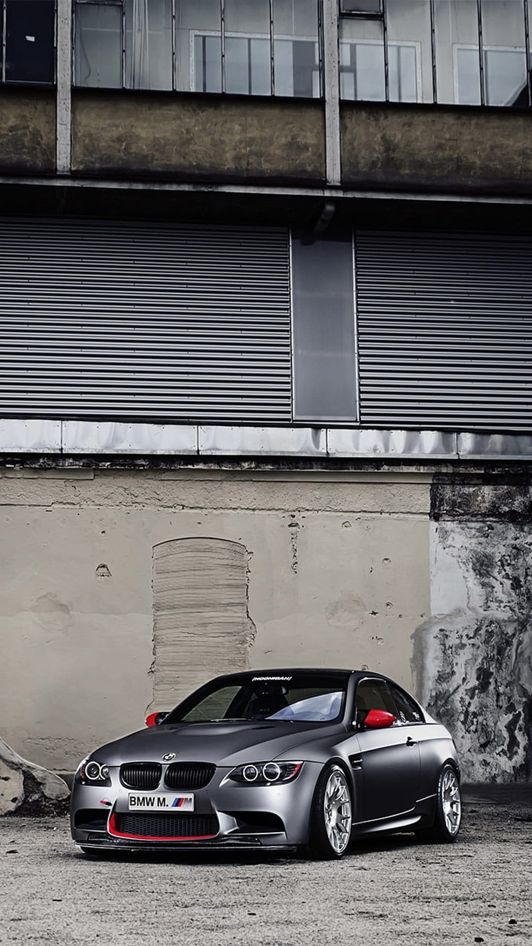 BMW M3 coupe in matte grey with red mirrors parked against a weathered industrial building with metal doors.