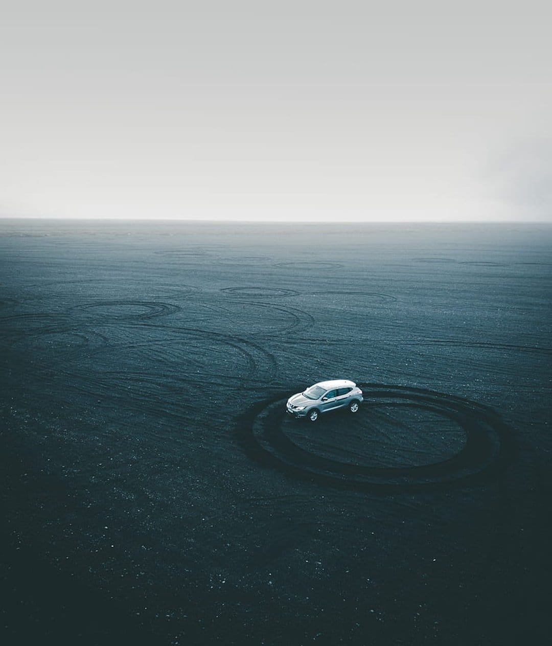 Silver SUV carves circular tire tracks on a black sand beach viewed from a high-angle aerial drone perspective.