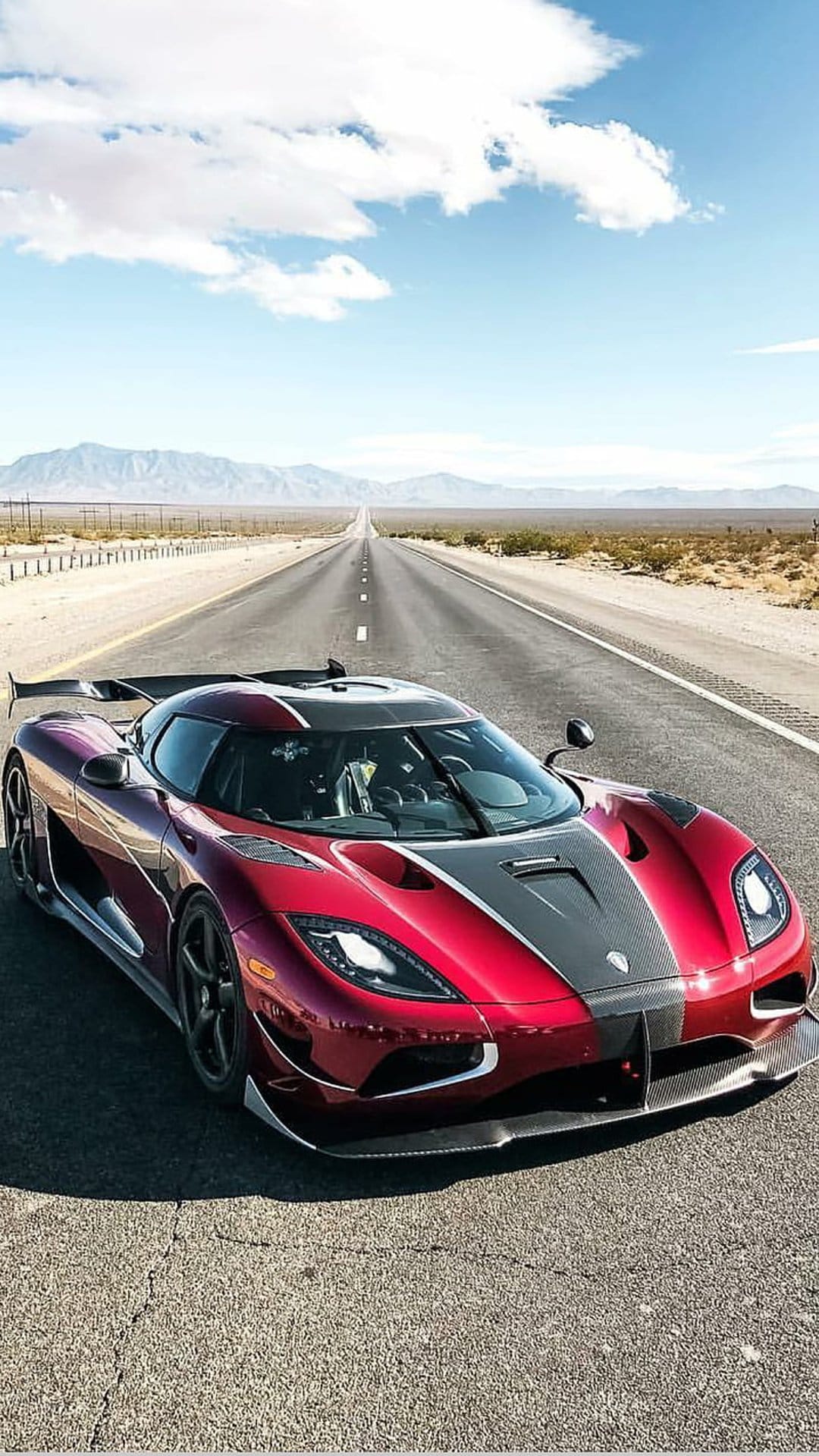 Red Koenigsegg Agera RS hypercar speeds along a sunlit desert road under a bright blue sky with white clouds.