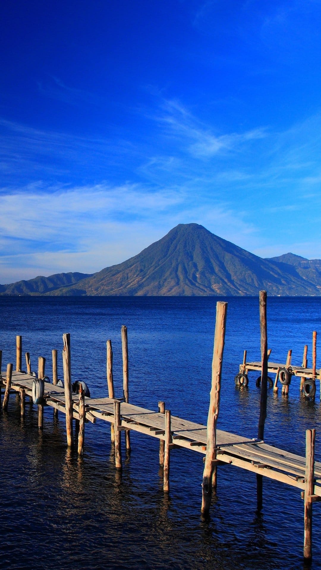 Weathered wooden pier leading into Lake Atitlan toward a massive volcano under a clear blue sky in Guatemala.