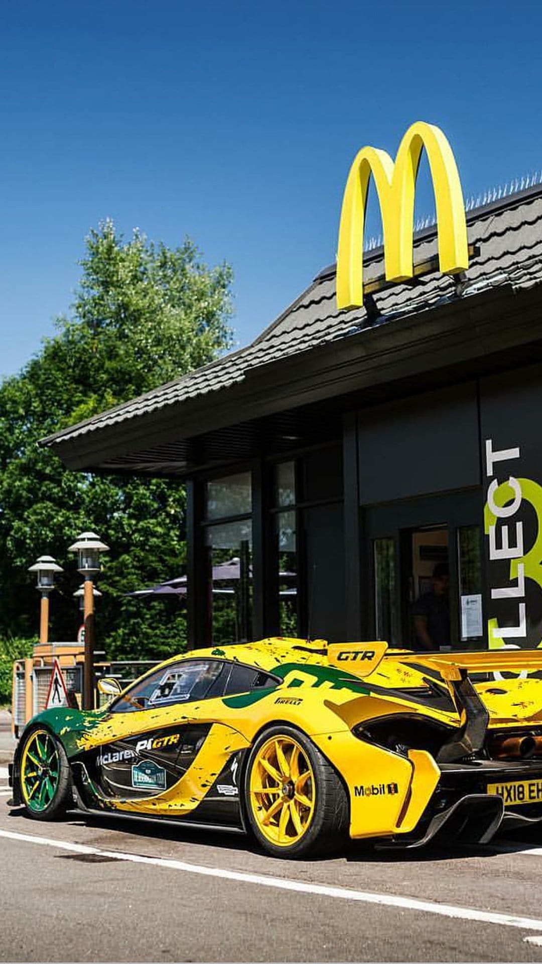Yellow McLaren P1 GTR racing car parked at a McDonald's drive-thru with green trees and a clear blue sky background.