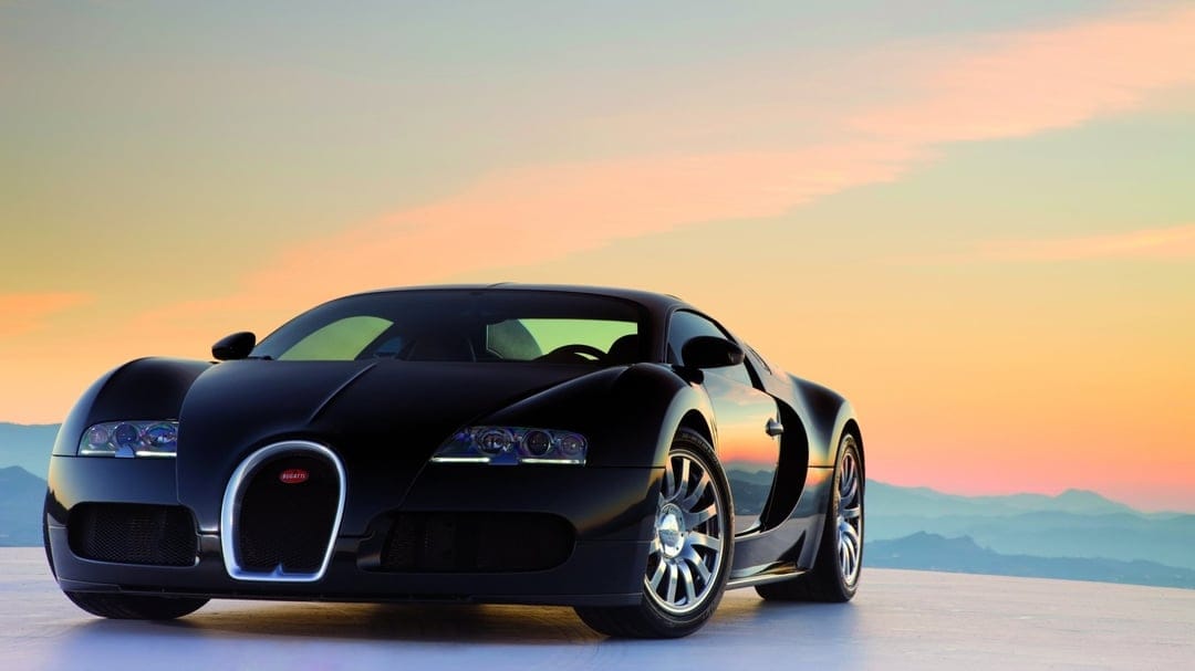 Black Bugatti Veyron supercar on a platform with a warm orange sunset and hazy blue mountains in the background.
