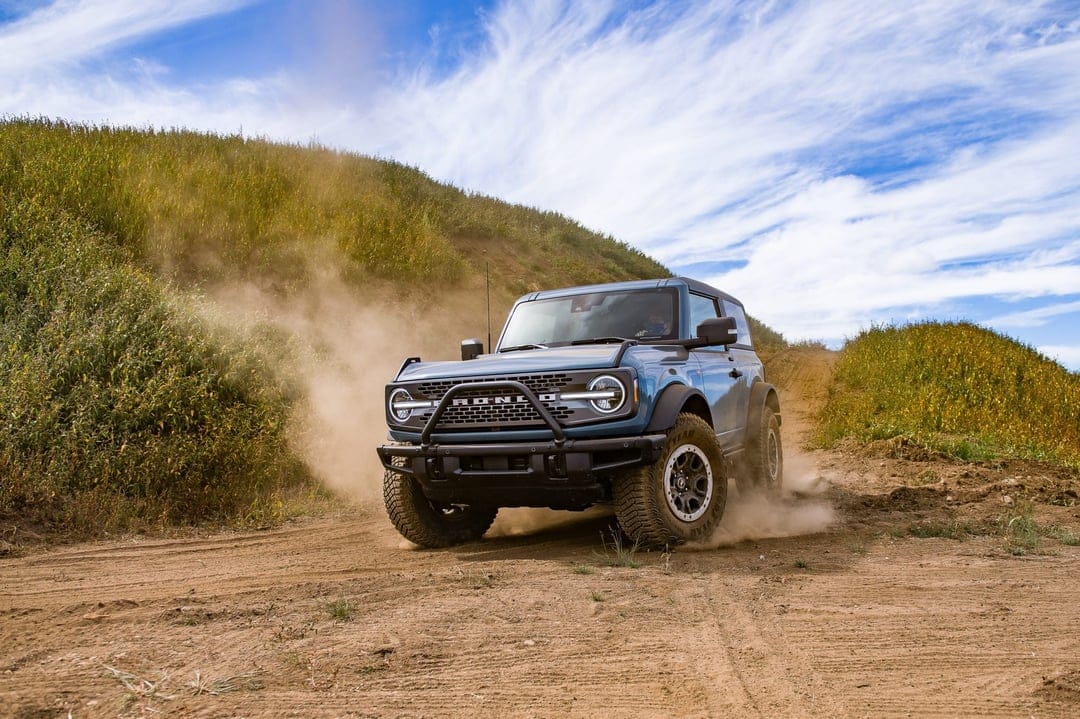 Blue Ford Bronco kicking up dust on a dirt trail between green hills under a bright sky with wispy clouds.