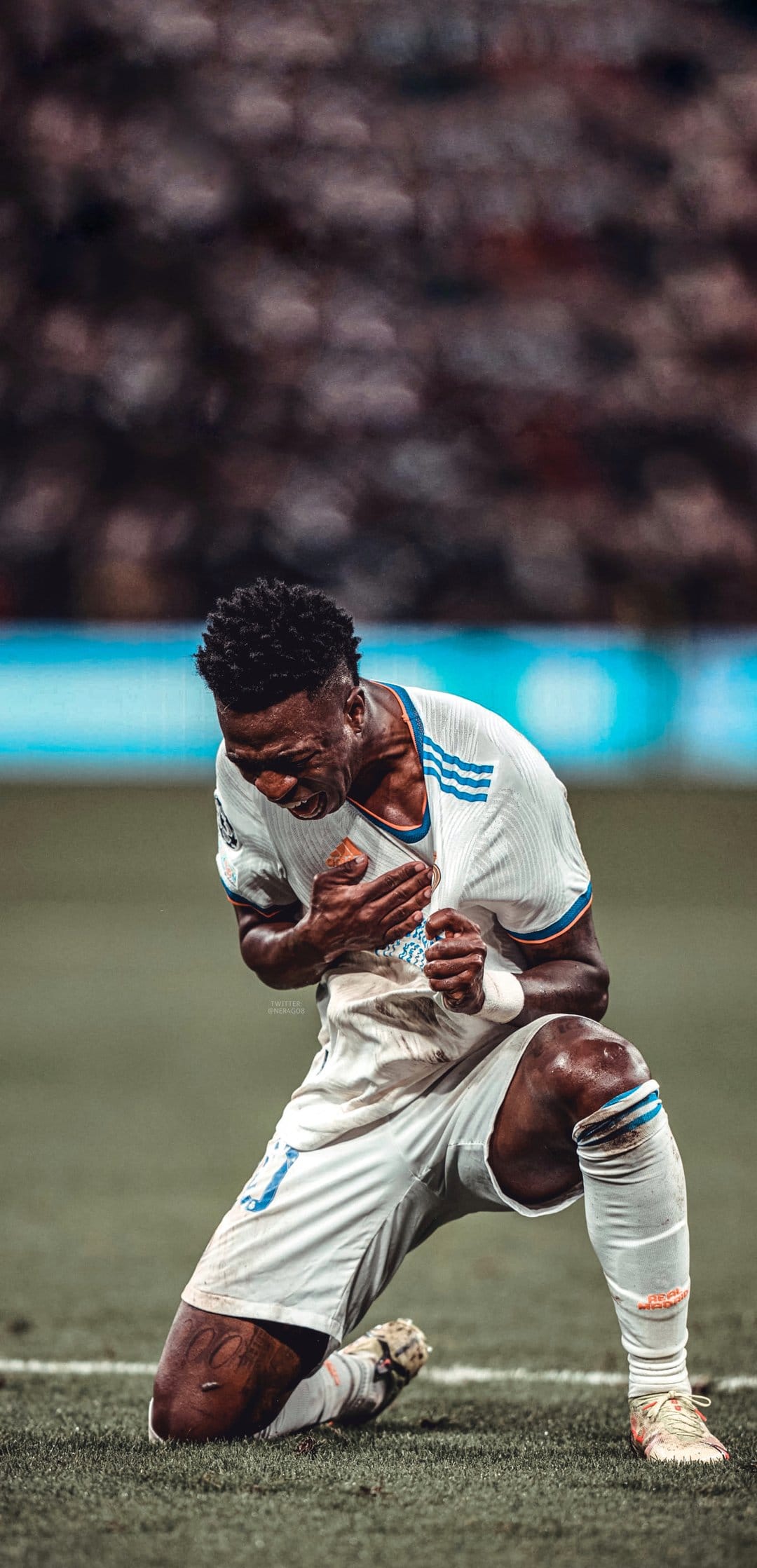 Vinicius Junior kneels on a green football pitch while clutching his white Real Madrid jersey in celebration.
