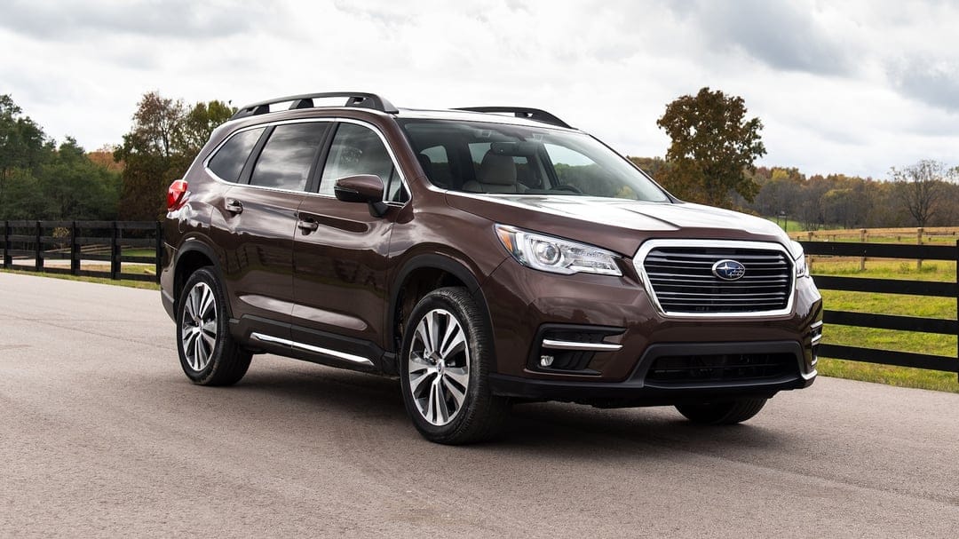 Brown Subaru Ascent SUV parked on a rural road beside a black wooden fence under a soft cloudy sky.