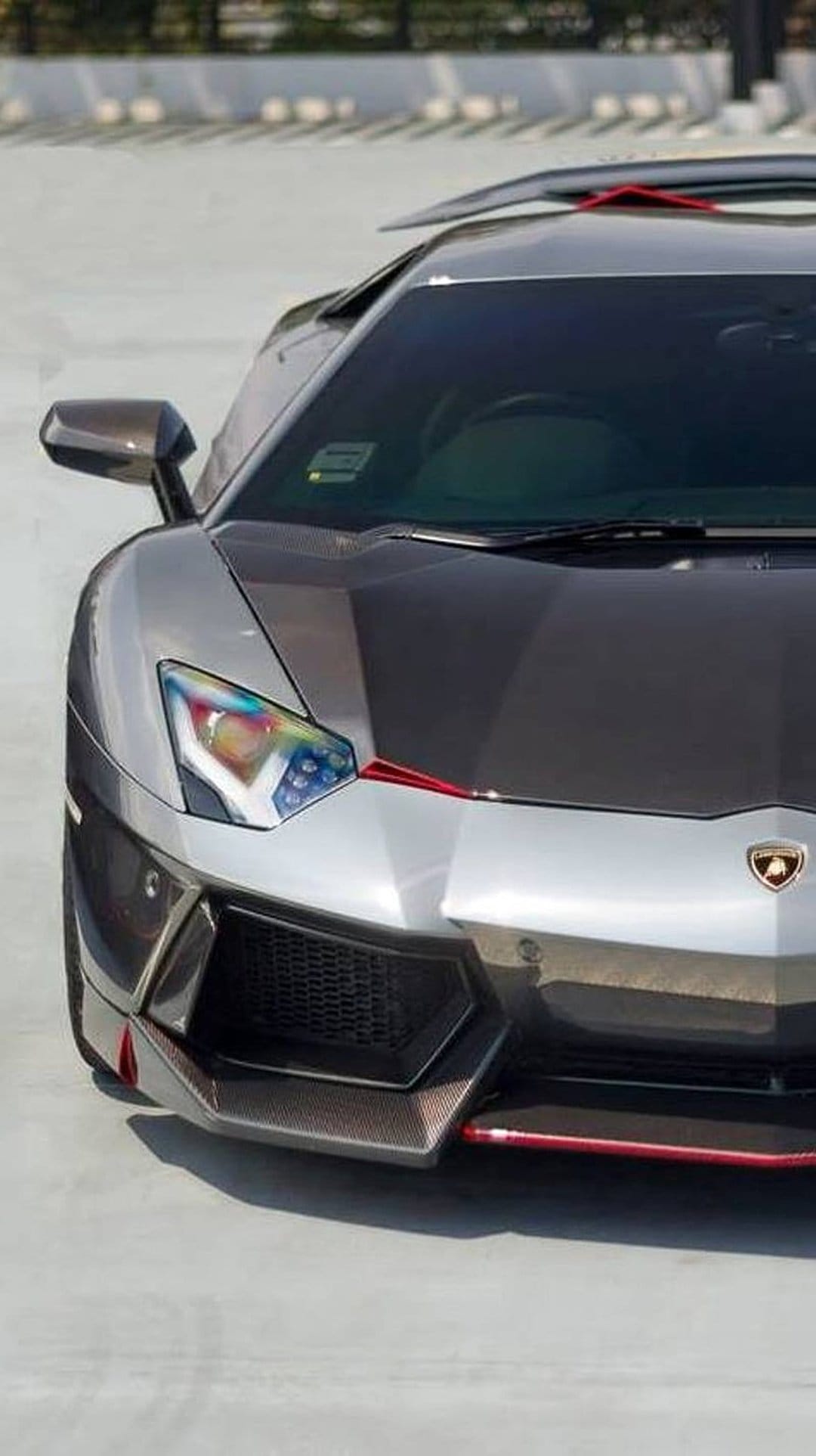 Silver Lamborghini Aventador with carbon fiber hood, rainbow headlights, and red accents on a concrete ground.