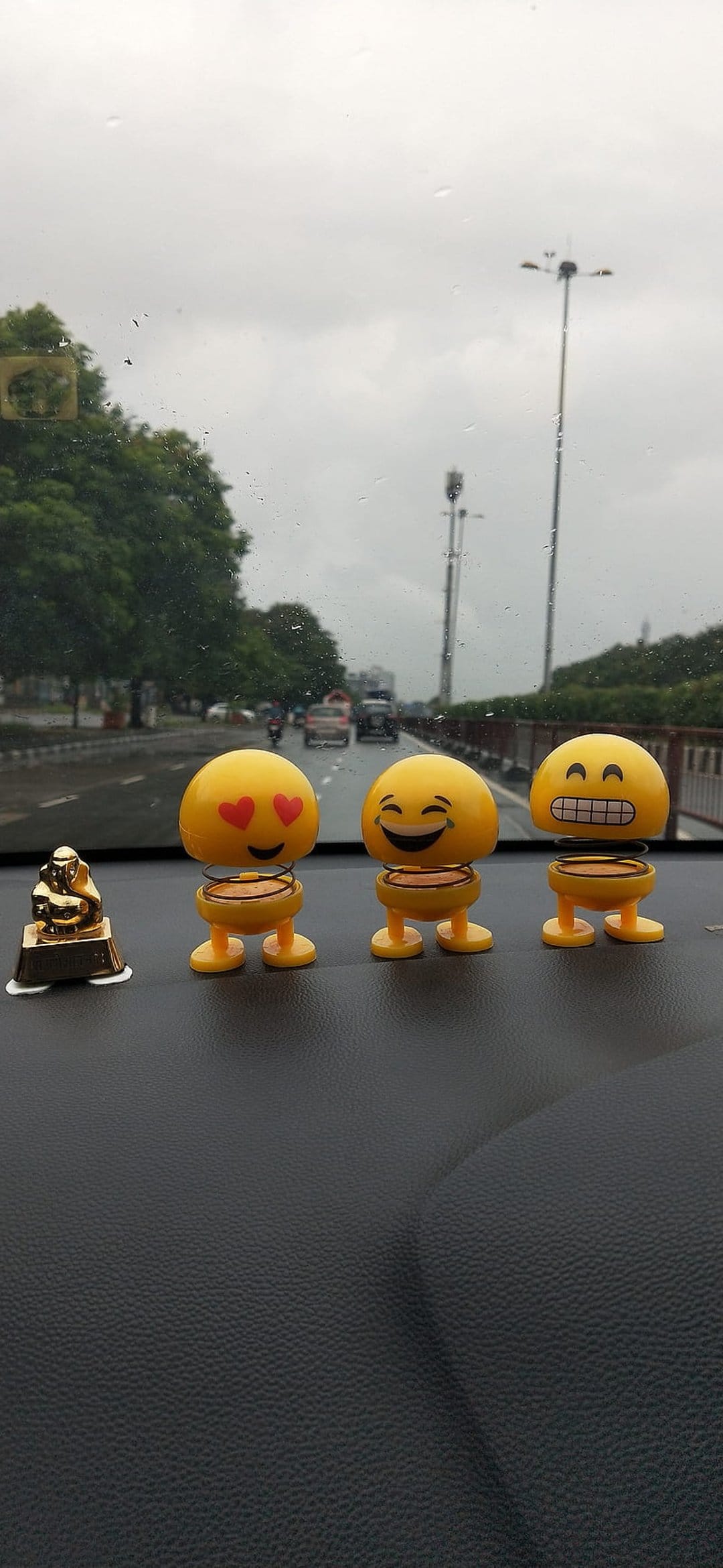 Yellow emoji bobbleheads and a golden Ganesha sit on a car dashboard against a blurred rainy street background.
