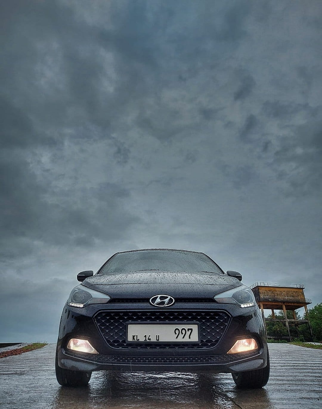 Black Hyundai i20 hatchback with headlights on parked on a wet street near a water tower under a cloudy sky.
