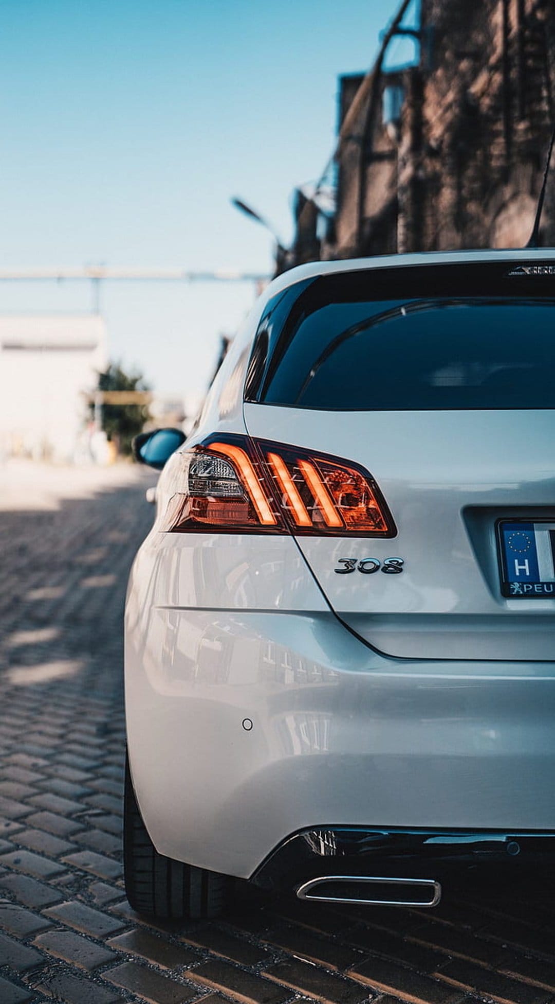 Peugeot 308 rear LED tail lights and 308 badge on a white car parked on a blurred urban cobblestone street.