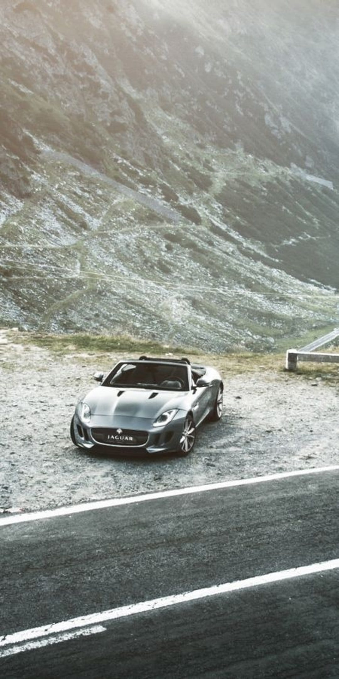 Silver Jaguar convertible parked on a gravel mountain road under bright sunlight with rocky cliffs behind it.