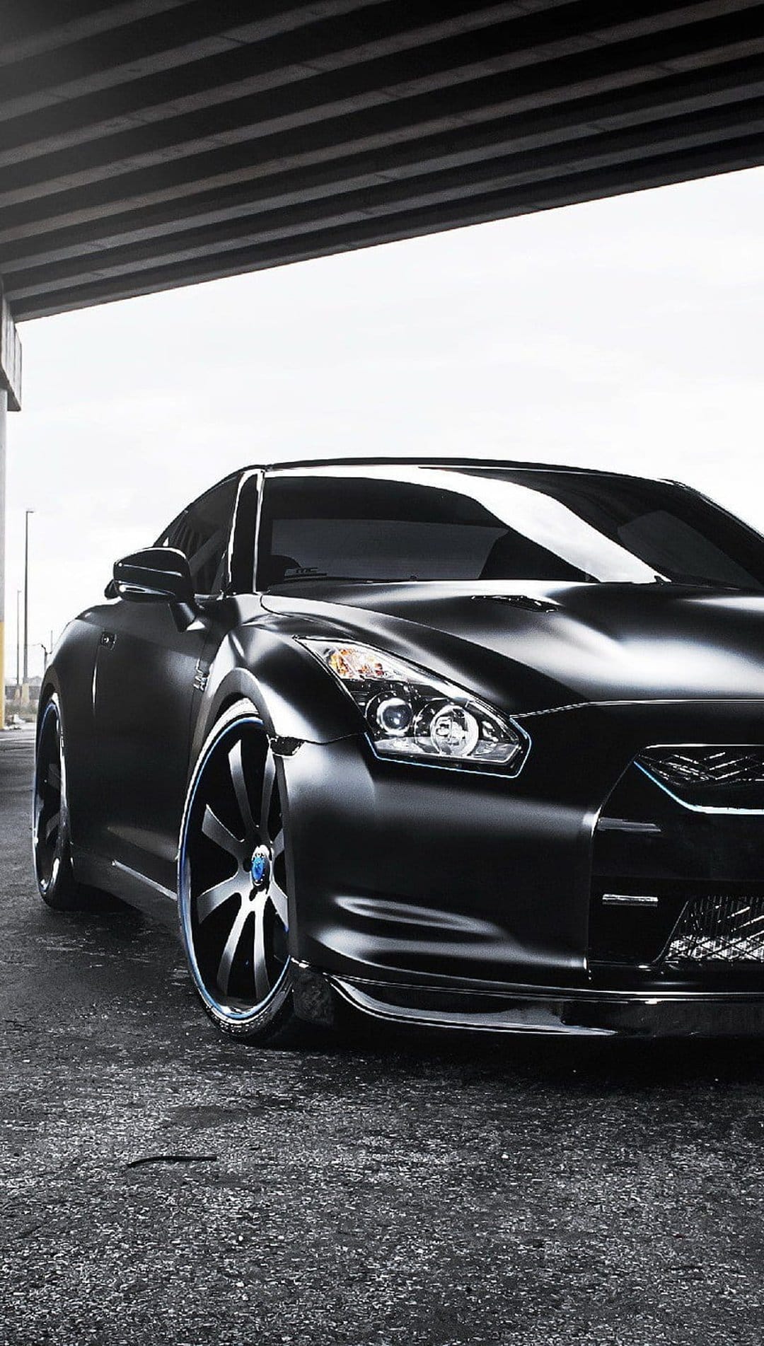 Nissan GT-R with blue wheel accents parked on asphalt under a concrete structure with reflections on black paint