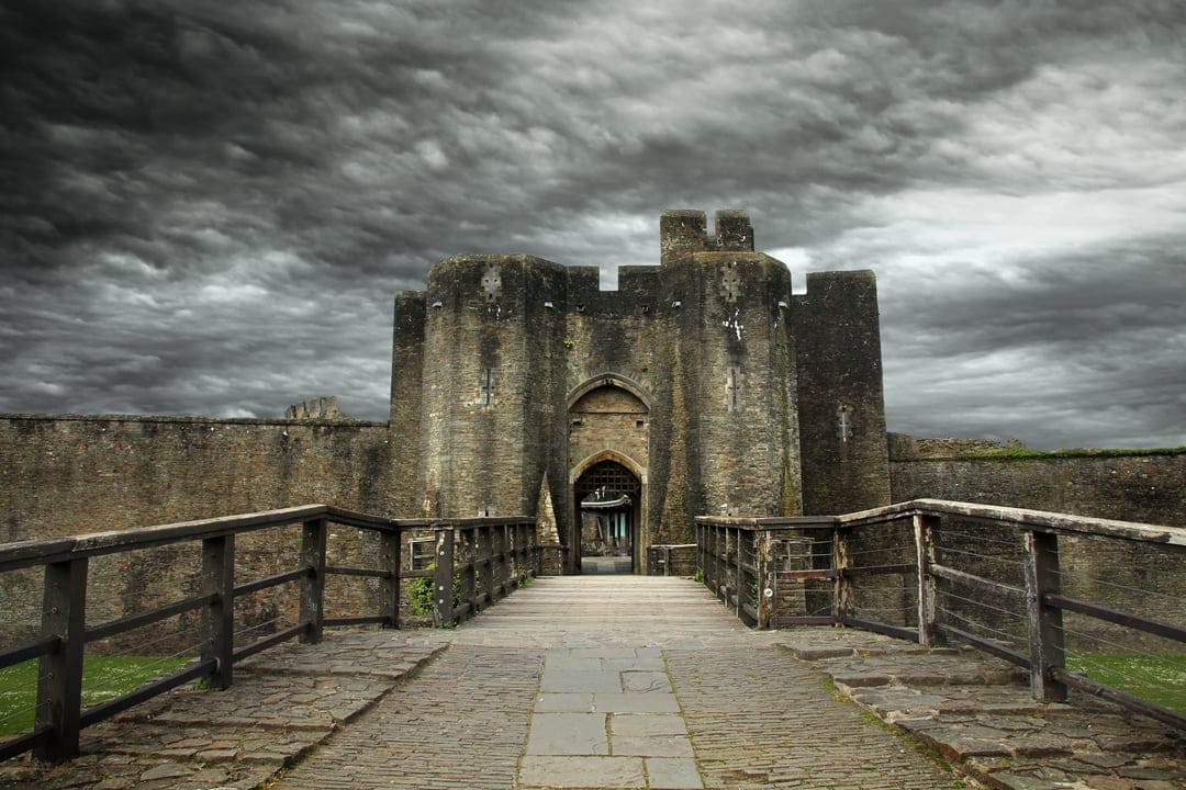 Ancient stone castle gatehouse with a wooden bridge stands under a dark, swirling sky of stormy grey clouds.