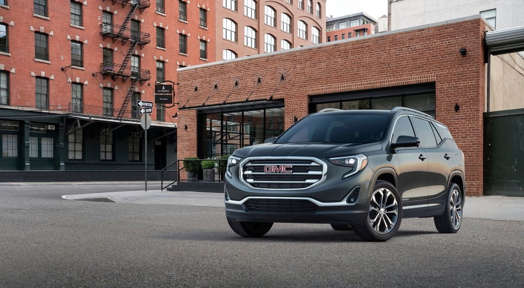 Dark grey GMC Terrain SUV parked on a city street in front of a multi-story red brick industrial building.