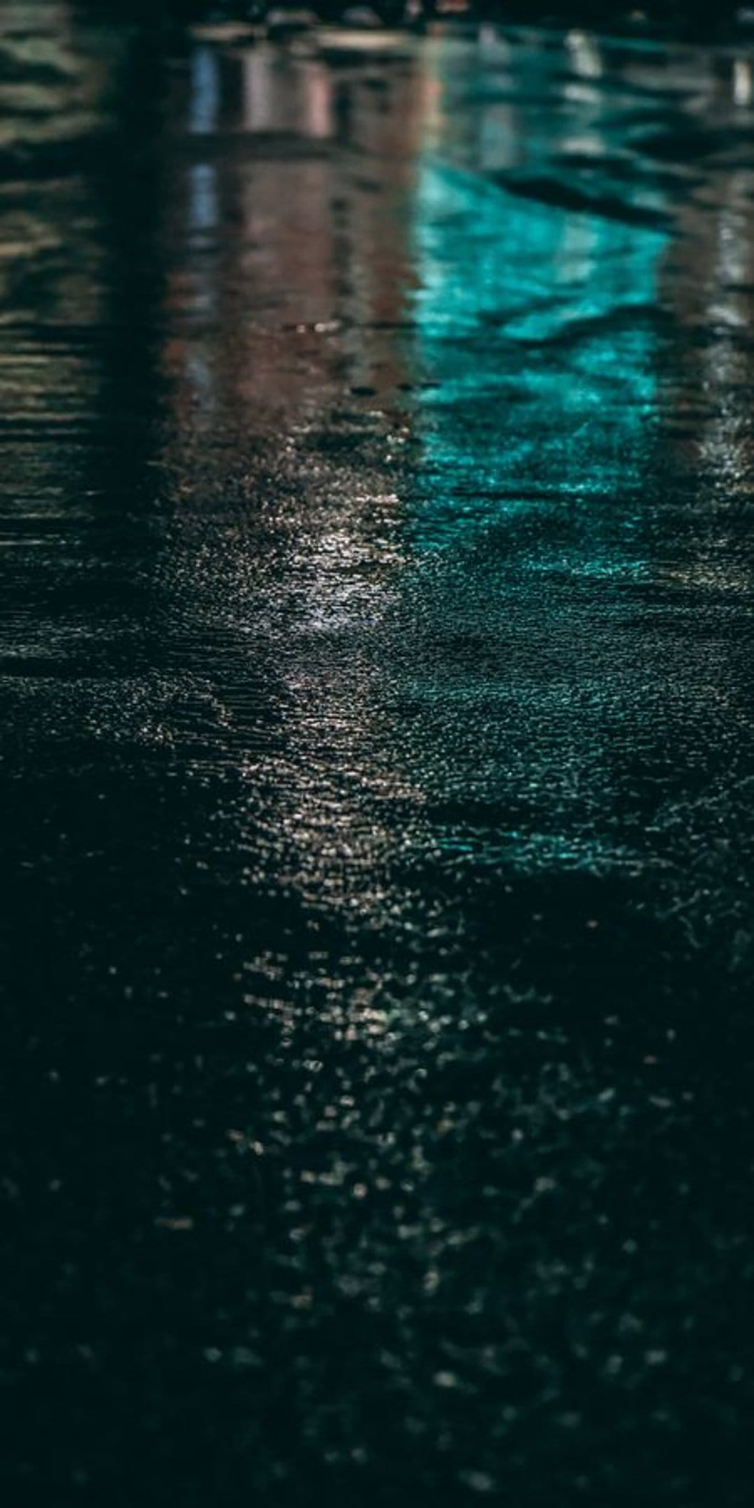 Teal and amber light reflections dance across a wet asphalt surface with soft ripples during a rainy urban night.