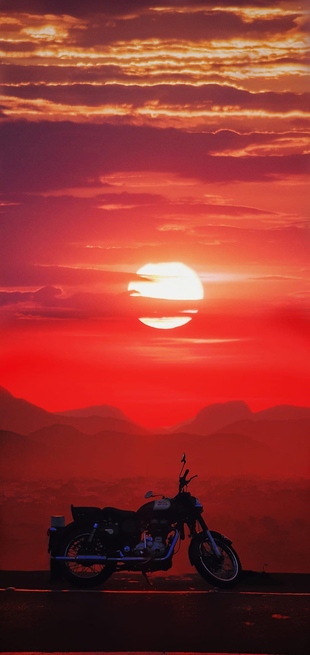 Motorcycle silhouette on a mountain ridge under a fiery red and orange sunset sky with distant layered peaks.