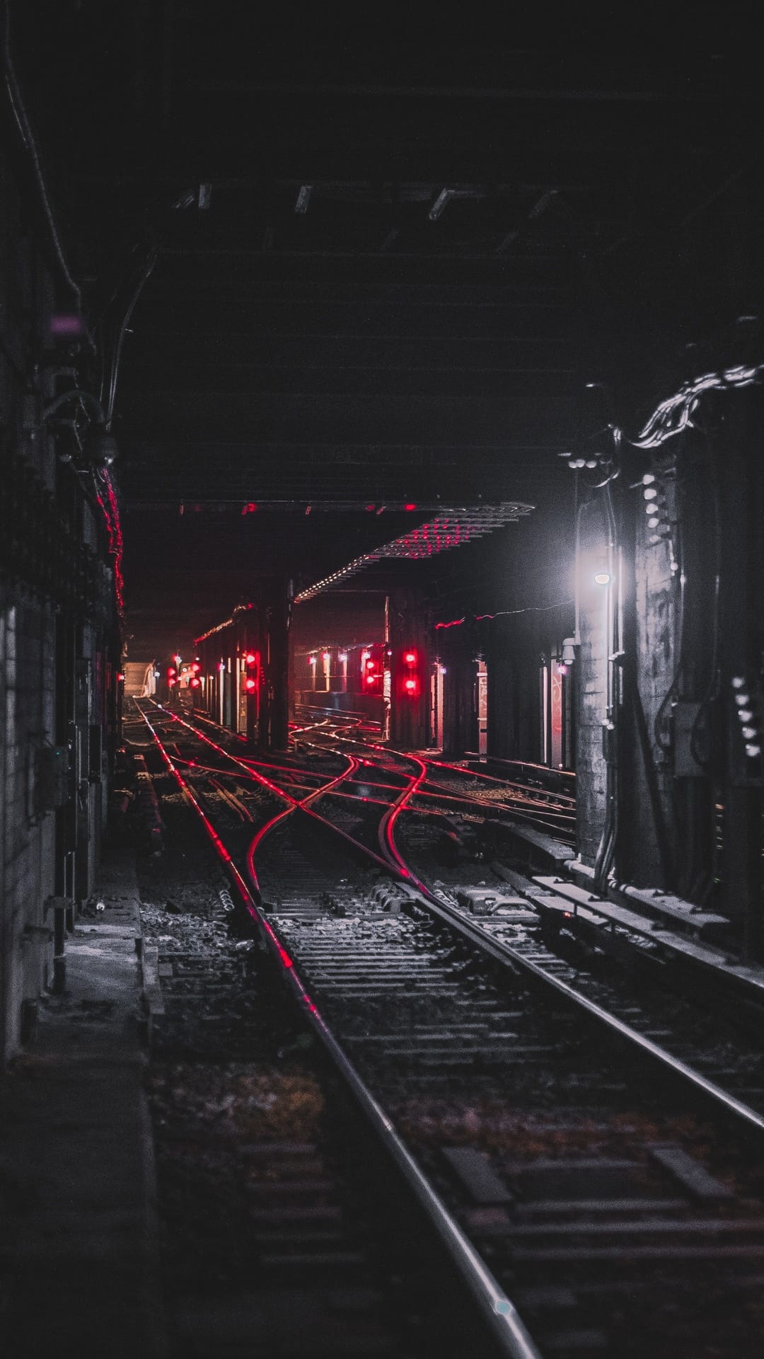 Subway tracks recede into a dark tunnel under glowing red signal lights reflecting on damp, industrial floors.