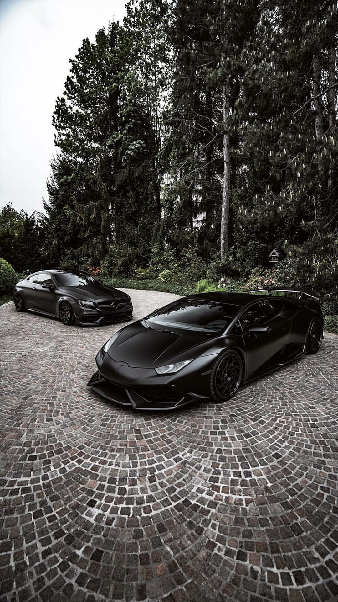 Matte black Lamborghini and Mercedes-Benz cars on a curved cobblestone driveway with dark trees in the background.