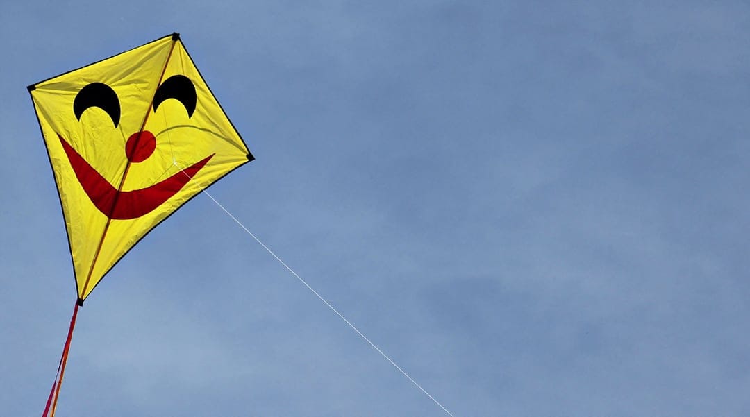 Yellow smiley face kite with a red grin soaring through a clear blue sky on a thin diagonal string tether.