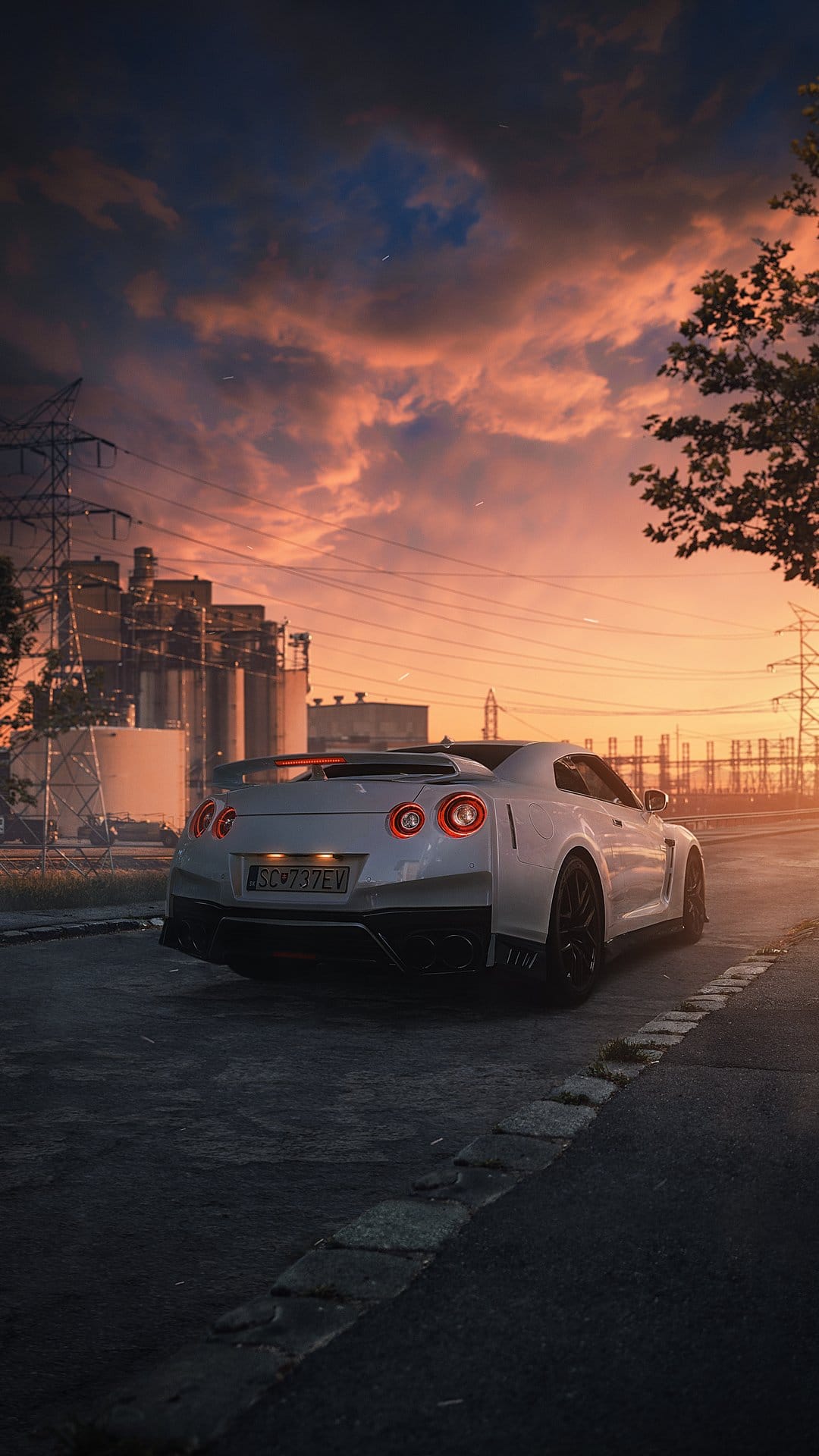White Nissan GT-R parked on asphalt with industrial power pylons silhouetted against a vibrant orange sunset.