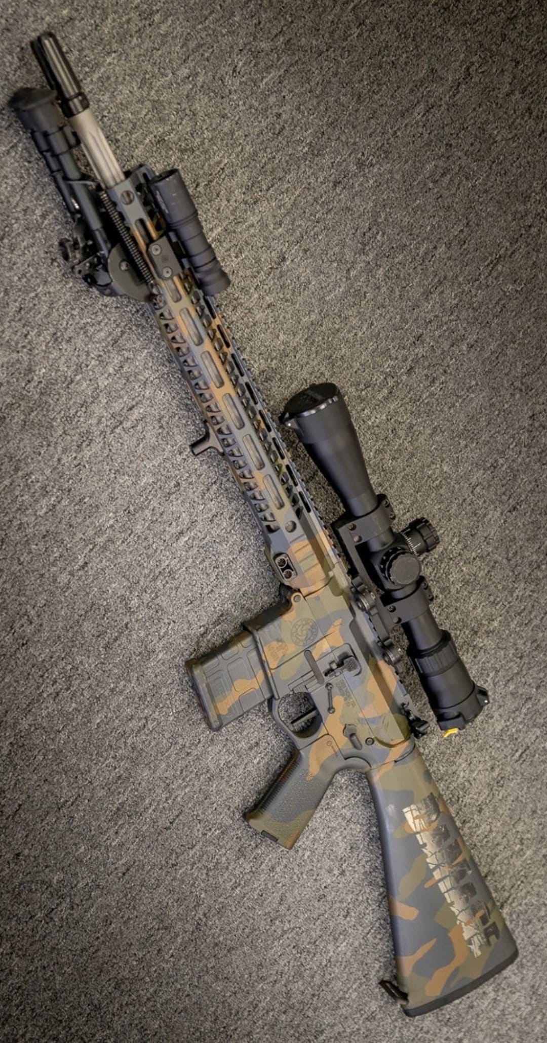 AR-15 rifle with brown and tan camouflage, magnified scope, and bipod resting on a flat surface in portrait view.