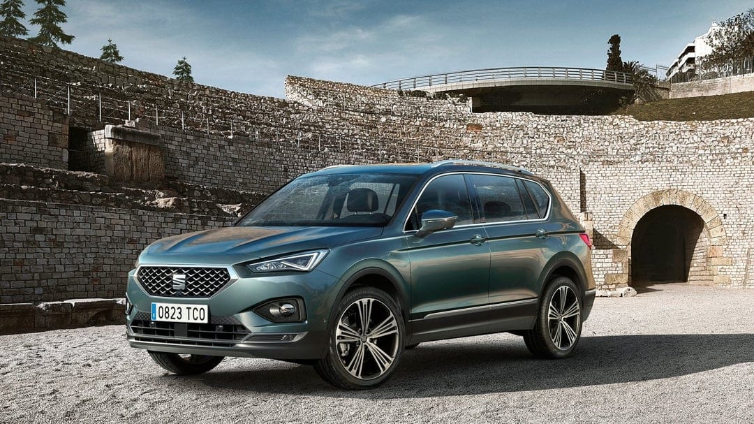 Dark teal SEAT Tarraco SUV parked on gravel in front of high ancient stone amphitheater walls under a clear sky.