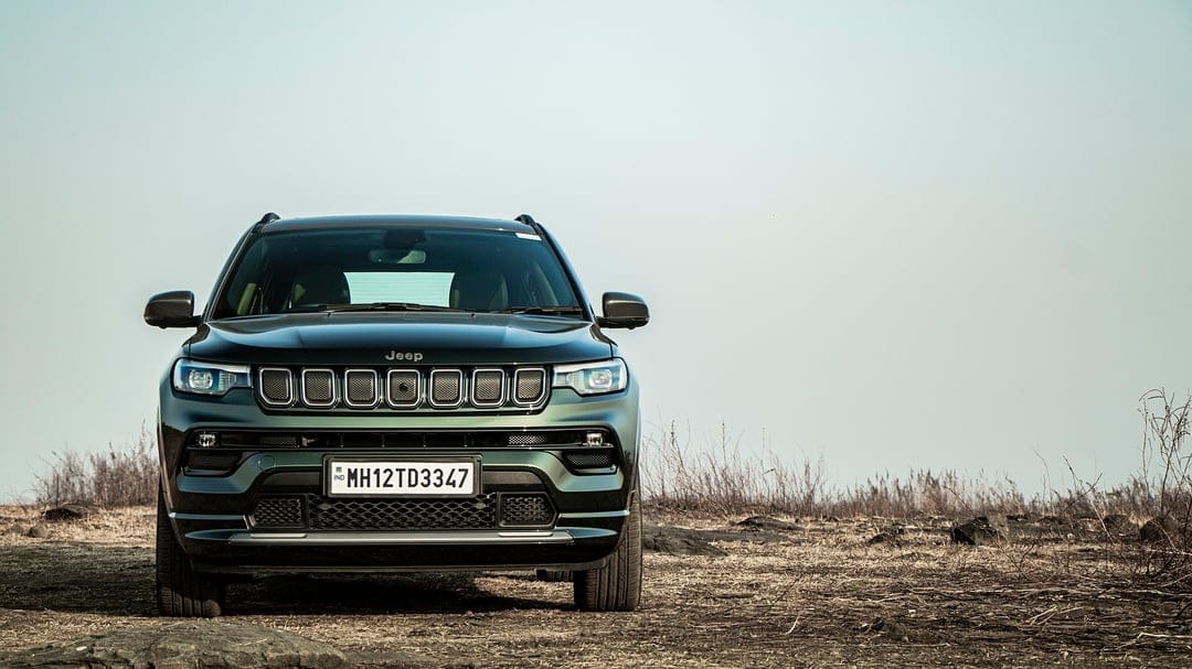 Dark green Jeep Compass SUV parked on a dry grassy field under a bright clear sky in a frontal shot.