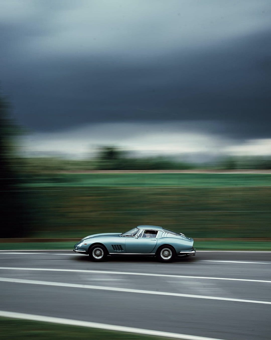 Blue Ferrari 275 GTB sports car racing on a grey road past blurred green trees under a dark, stormy sky.