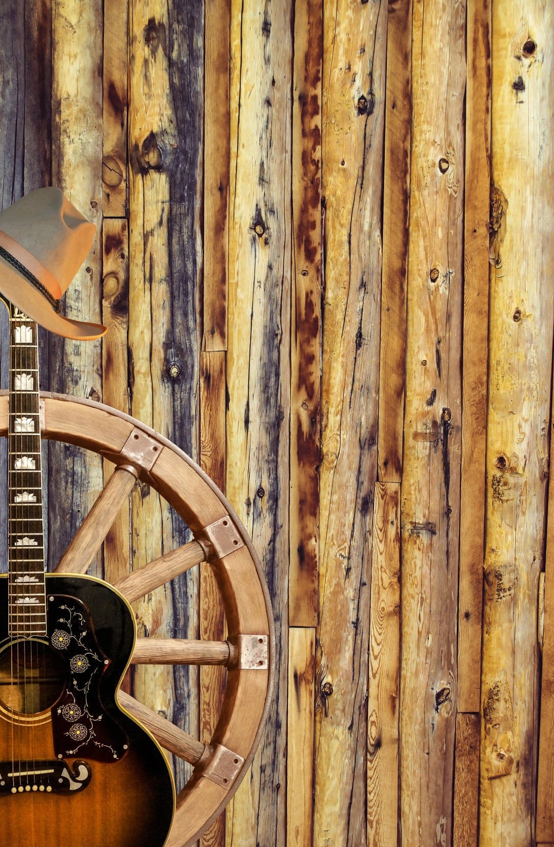 Acoustic guitar rests against a weathered barn wall beside an antique wooden wagon wheel in a rustic setting.