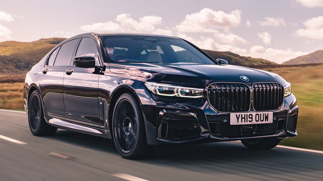 Black BMW sedan driving on a winding road with green hills and a soft cloudy sky in the background.