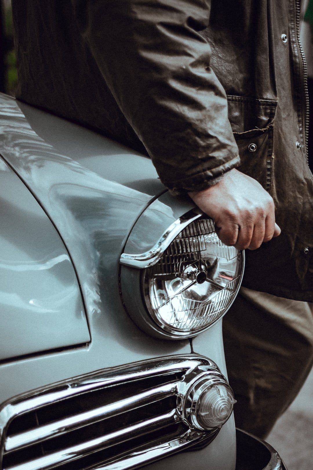 Man's hand in a dark jacket rests on the circular chrome headlight of a classic light blue vintage automobile.