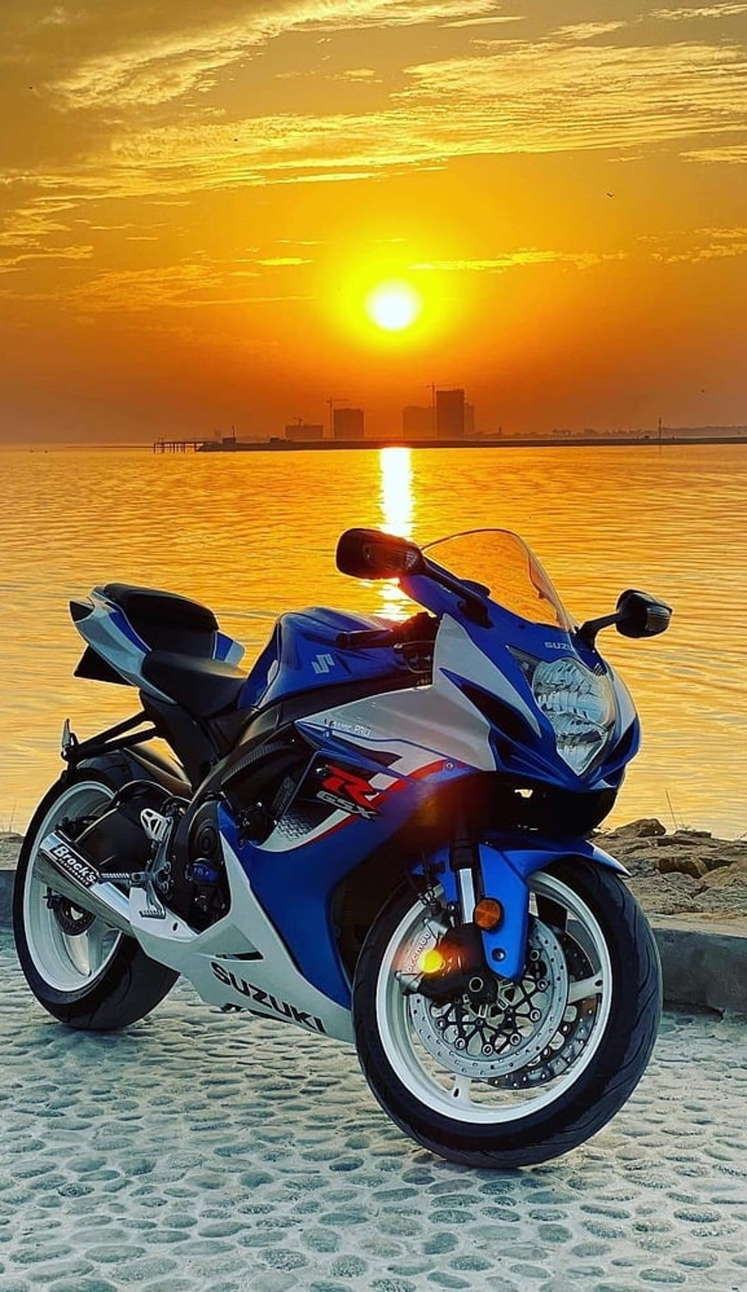 Suzuki sportbike in blue and white on a cobblestone beach during a golden sunset with a distant city skyline.