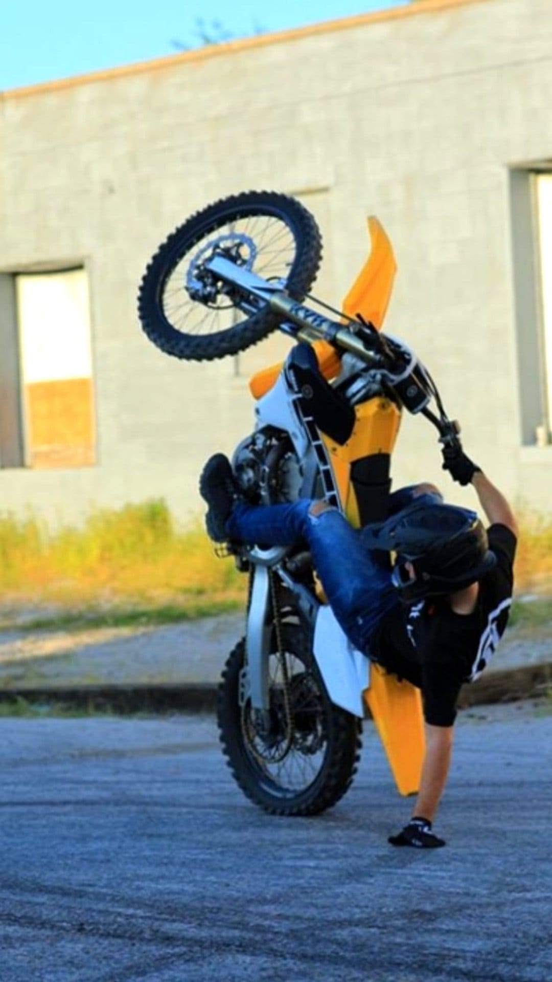 Motorcyclist in a helmet executes a steep wheelie on a dirt bike against a blurred field and building.