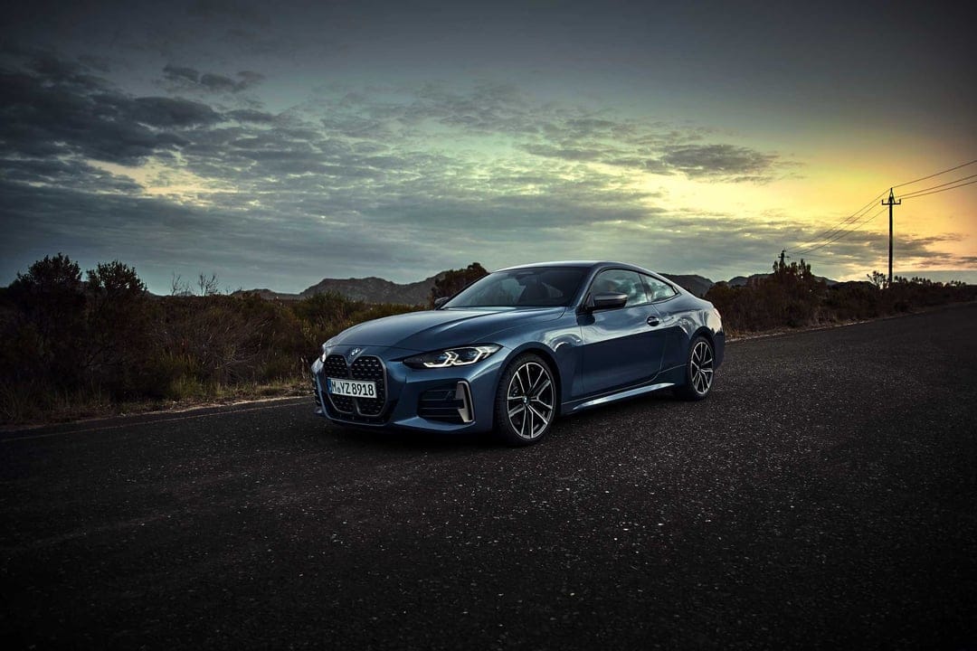 Blue BMW 4 Series Coupe on an asphalt road at dusk with hills and a sunset sky in the background.