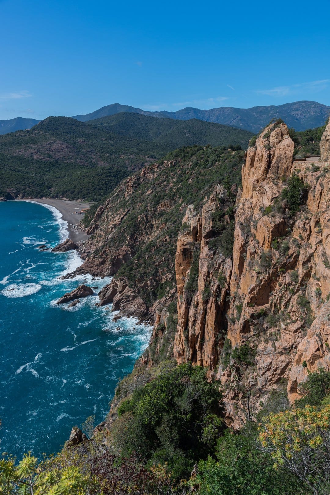 Orange coastal cliffs rise above deep blue Mediterranean waves and a pebble beach under a clear summer sky.