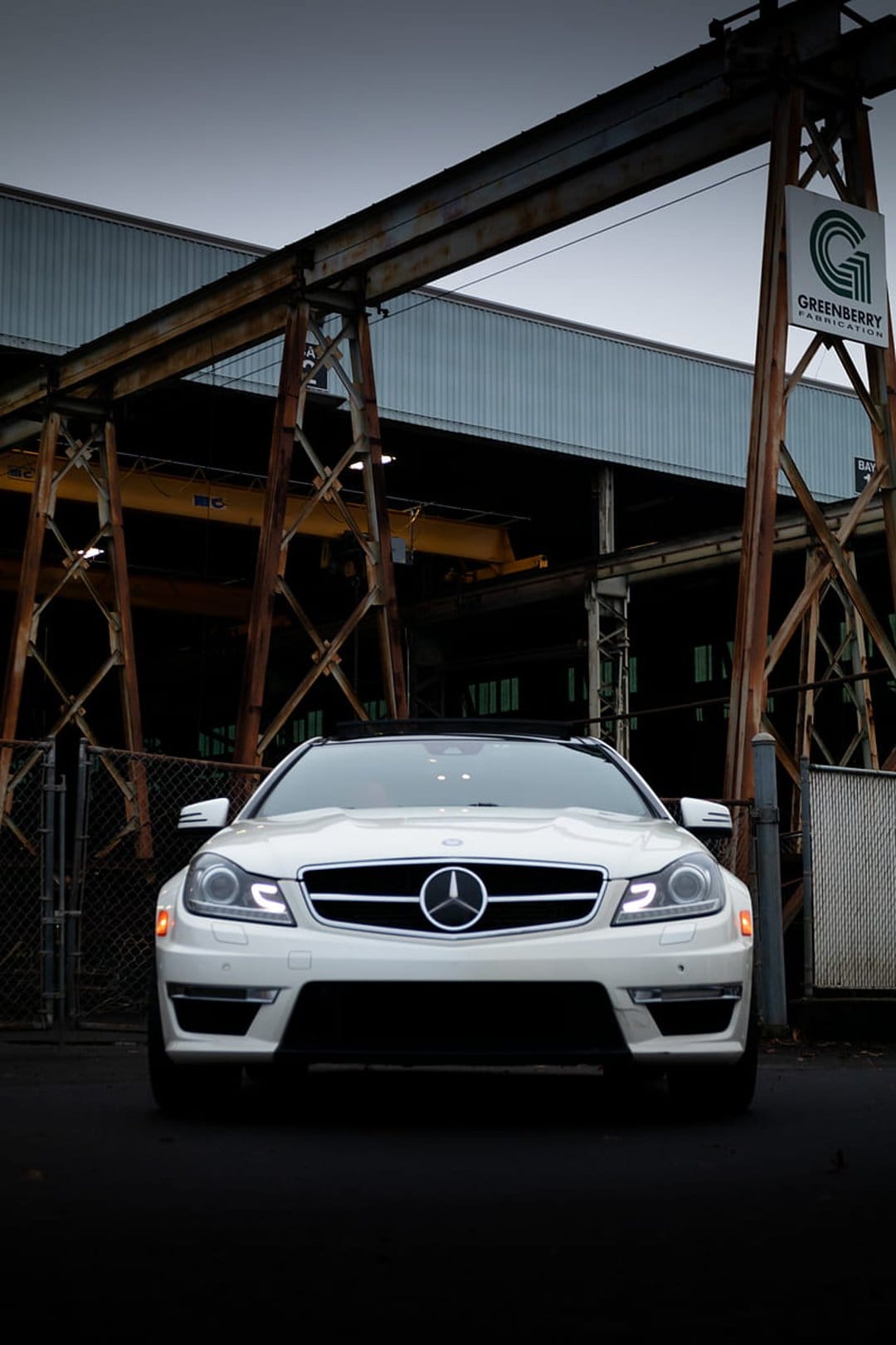 White Mercedes-Benz C-Class coupe with glowing headlights parked in front of a dark, rusted industrial factory wall.