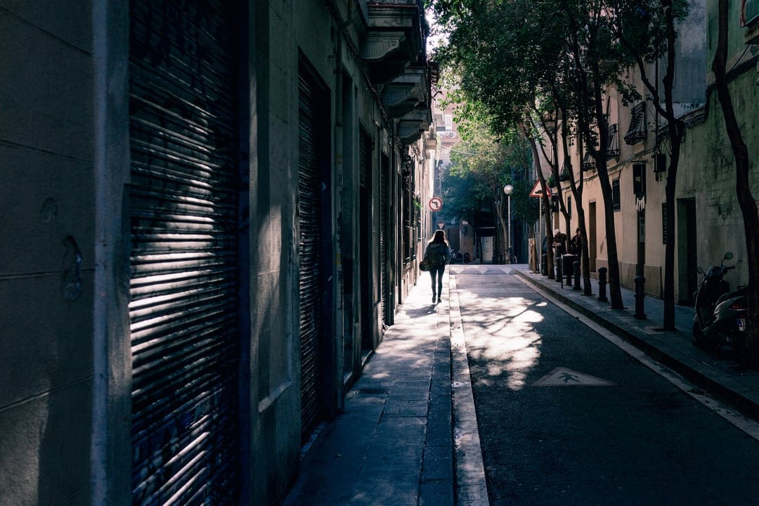 A woman walks through a sun-dappled European alleyway with cobblestone floors and tree-shaded historic buildings.