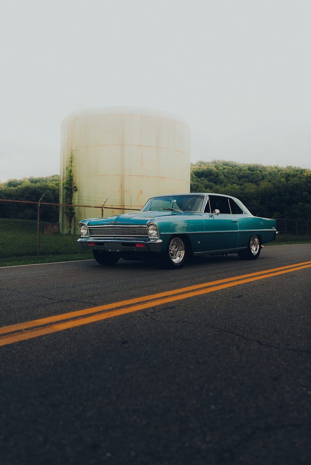 Teal Chevrolet Nova sits on a road beside a large weathered industrial tank under a cloudy sky with green trees.