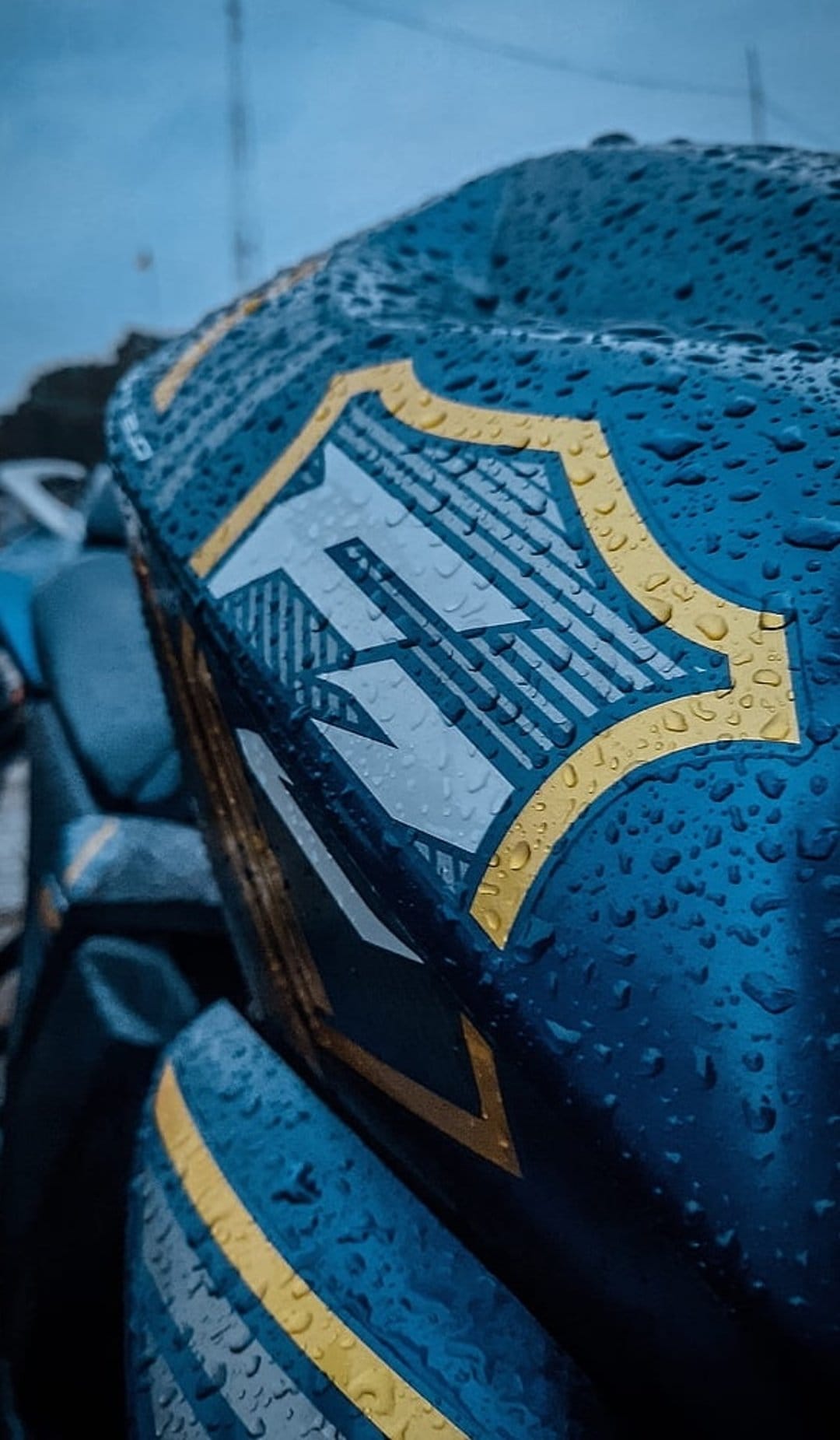 Blue and gold shield emblem featuring a striped letter M covered in tiny raindrops against a moody sky backdrop.