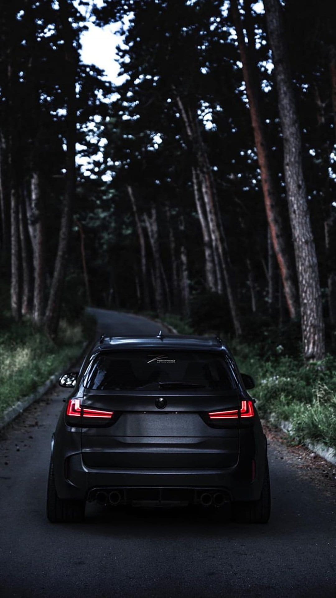 Matte black SUV with glowing red taillights driving through a dark, dense forest on a winding asphalt road.