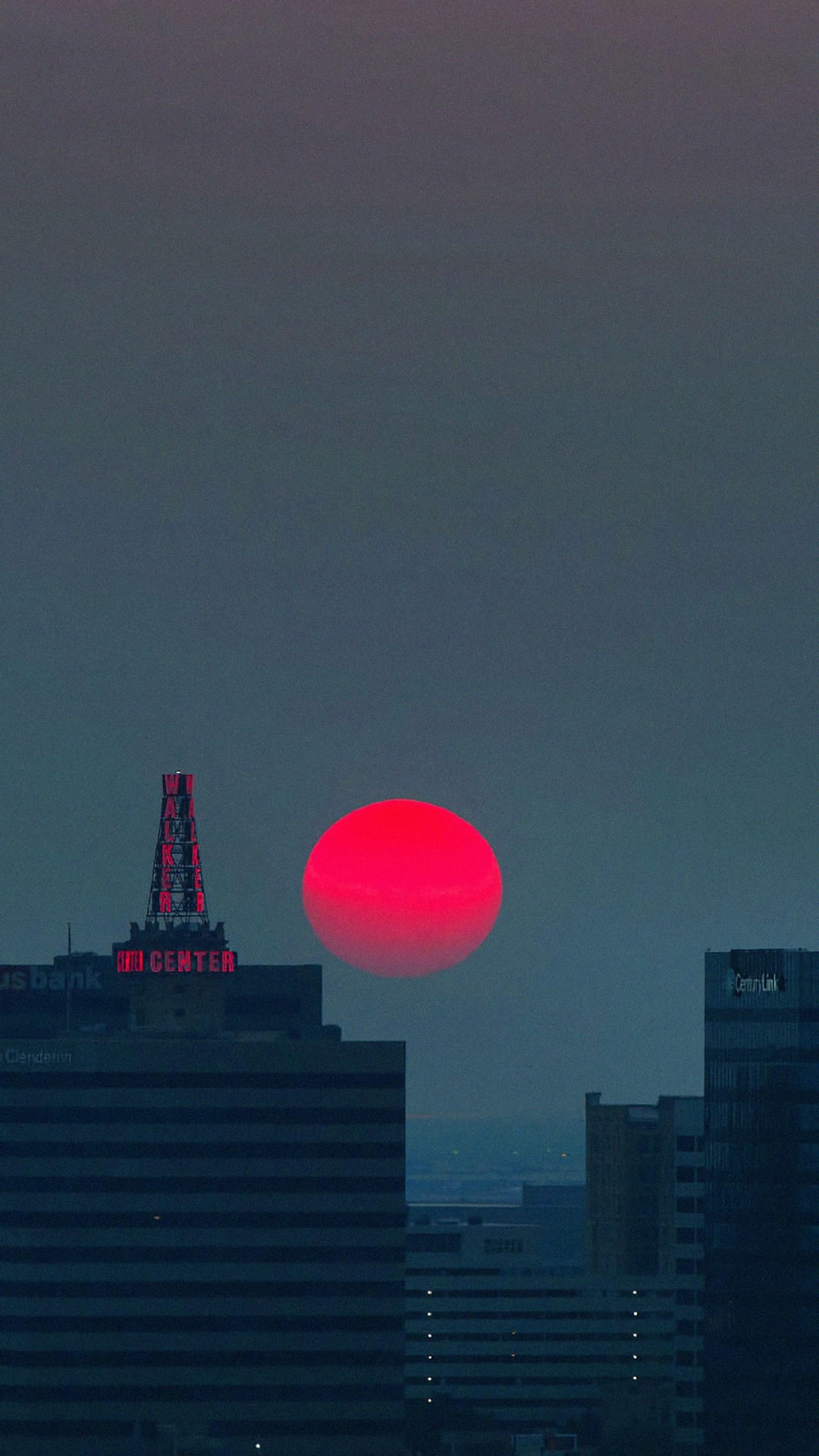 Large red sun descending behind a dark urban skyline silhouette during a dramatic twilight transition.