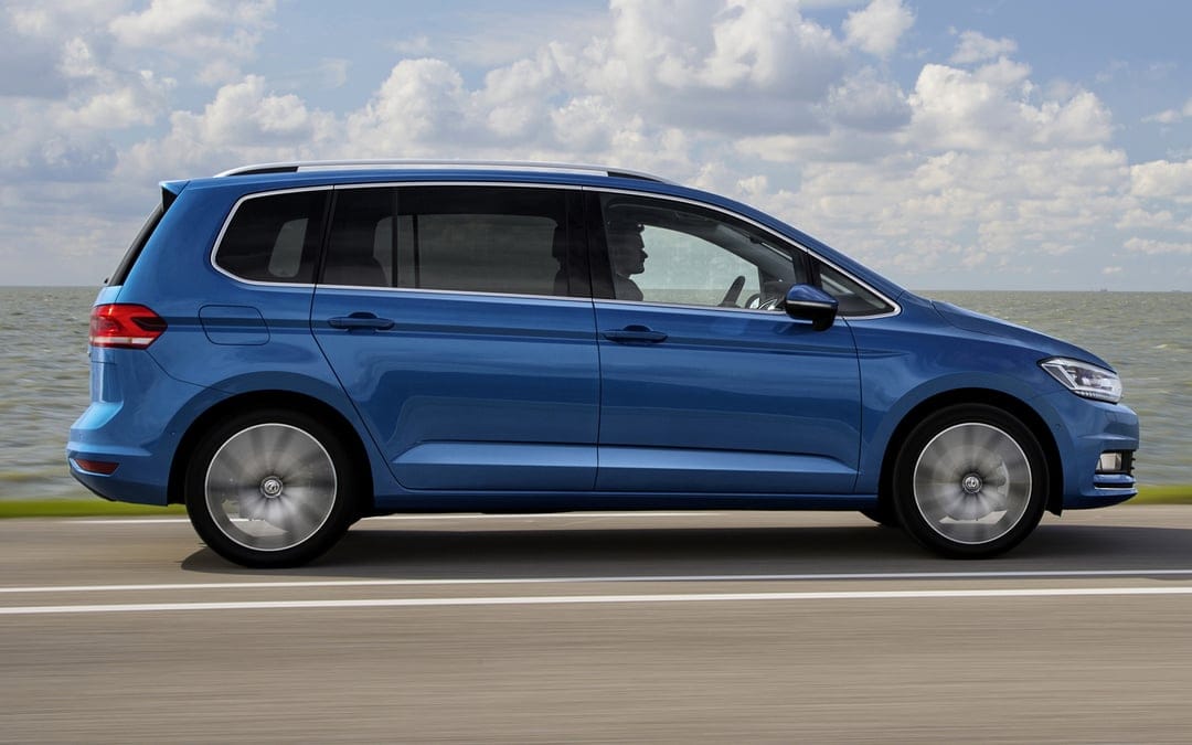 Blue Volkswagen Touran driving on a coastal road with motion-blurred wheels under a bright, cloudy sky.